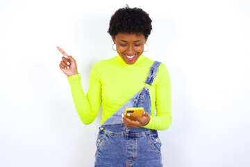 Smiling African American female with curly bushy wears jeans overalls over white wall pointing finger at blank space holding phone in one hand