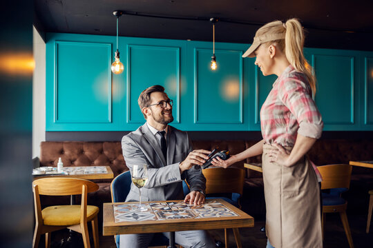 Online payment of restaurant bills. Man in a business suit with a tie sits at a table in a restaurant and leans his phone on the terminal held by a female waitress. Paying for a meal during lunch time