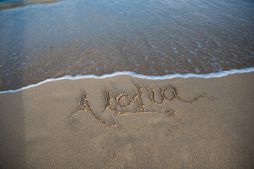 The word Aloha is written into the sand as the surf washes on shore at the beach.