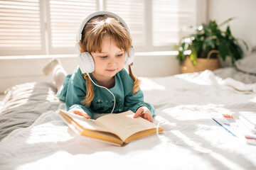 Funny girl in a blue dress reading a book on a white bed in a sunny bedroom