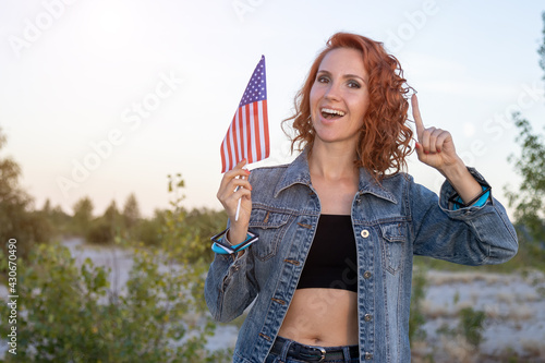 Young happy patriotic woman with the US flag in nature. 4th of july independence day of the united states. Beautiful red-hair vivacious girl with the American flag standing in the summer sunshine day.