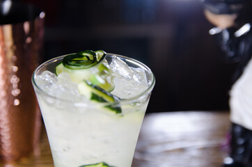 white cocktail with cucumber and ice in a bar at night, party invitation template with free space for text