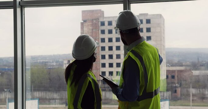 Two construction engineers standing indoors near big window looking at the unfinished concrete building in front, discussing future real estate project.