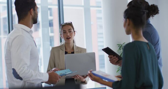 Caucasian businesswoman holding laptop talking to diverse colleagues standing at office meeting