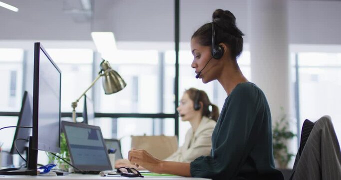 Mixed race businesswoman sitting at desk talking using phone headset and computer in busy office