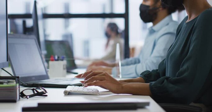 Diverse male and female colleague in face masks using computers at desks separated by sneeze shield