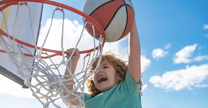 Close up image of kid basketball player making slam dunk during basketball game in floodlight basketball court. The child player is wearing sport clothes.