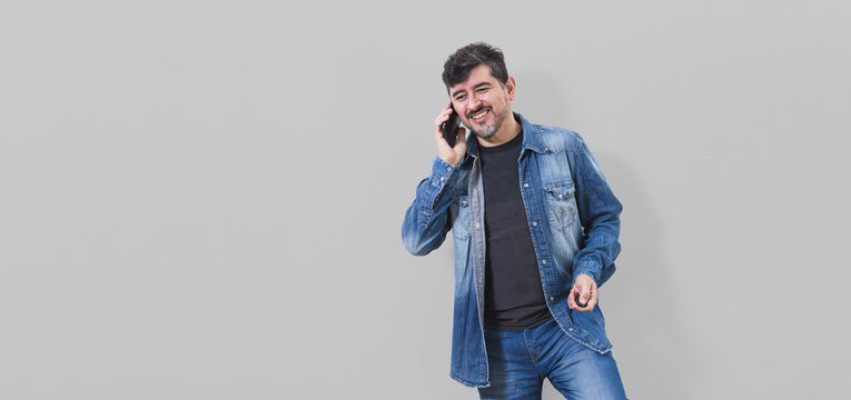 Panorámica de hombre latino muy feliz y sonriente hablando por teléfono vestido con camisa de denim y remera negra