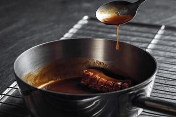 Process of pouring sauce over octopus in silver cooking pot from silver spoon