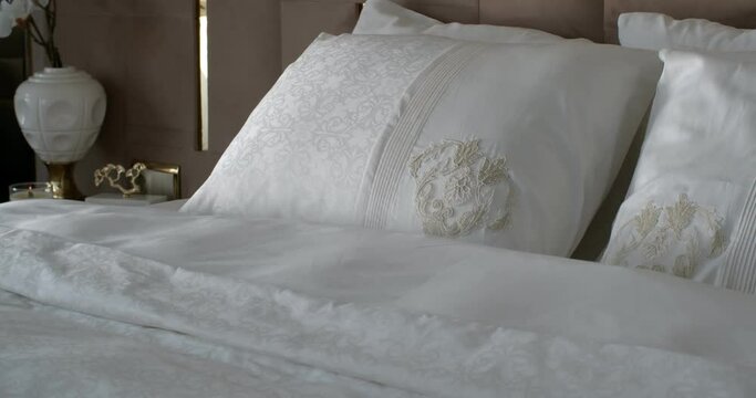 White linens with floral patterns on the large double bed. The wall behind the bed is upholstered in soft gray material. There is a bedside table with an artificial plant in a white vase and a scented