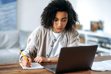 Attractive successful smart young African American woman in stylish clothes, freelancer or student working or studying remotely, sitting in living room at laptop, looking at screen, taking notes