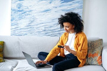 Online shopping concept. Young beautiful african american woman in casual stylish clothes sits on the couch, makes purchases online by laptop, fills in credit card details to pay for goods or delivery - obrazy, fototapety, plakaty