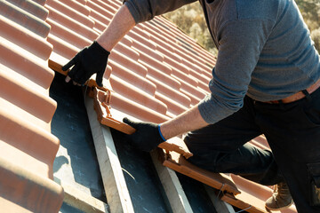 Closeup of worker hands installing yellow ceramic roofing tiles mounted on wooden boards covering residential building roof under construction. © bilanol