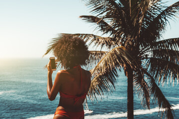 View from behind of a slim young African-American female in a swimsuit using her smartphone and...