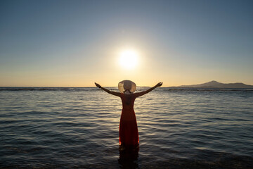 Young woman wearing long red dress and straw hat raising hands standing in sea water at the beach enjoying view of rising sun in early summer morning.