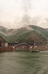 Foto op Canvas Baidicheng, China - May 7, 2010: Qutang Gorge on Yangtze River. Brown concrete road bridge along shore over sanded inham set in a green mountain landscape with barge in front. foggy cloudscape. © Klodien