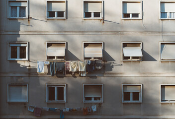 A greyish house facade of a European residential house in uptown with many windows, part of which is covered by external blinds, with clotheslines with drying clothes on it, long sunset shadows