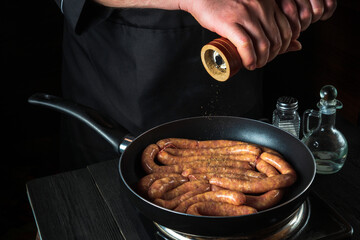 The chef adds pepper to pan with raw meat sausage. Preparation for cooking sausages in kitchen of a restaurant or cafe on table with vegetables and spices