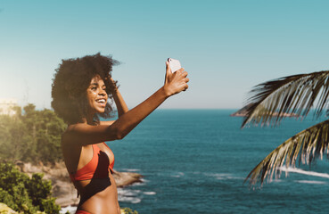 A funny ravishing laughing young Brazilian female is having a video vlog conversation via her smartphone or making a selfie while standing in a red swimsuit in front of the ocean, in a tropical resort