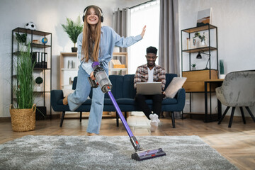 Smiling woman in headphones using hand held vacuum cleaner for chores while her afro american man...