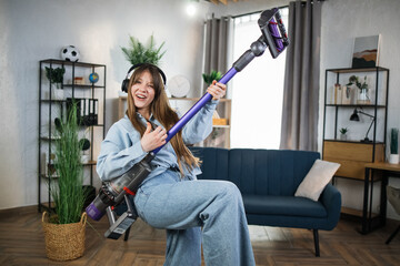 Positive young woman listening music in wireless headphones and playing with handheld vacuum...