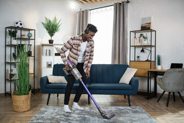 Smiling afro american man in wireless headphones cleaning carpet with handheld vacuum cleaner. Young guy enjoying housekeeping using modern devices. - obrazy, fototapety, plakaty