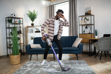 Playful african guy in casual wear removing dust from carpet with handheld vacuum cleaner. Happy...