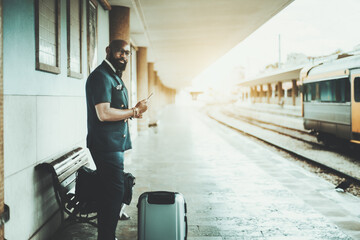 Fototapeta na wymiar A masculine bald bearded black man entrepreneur is using a smartphone while waiting for a high-speed railway train on a railroad station platform with two bags of luggage to start his business trip