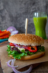 Freshly made burger on the cutting board on the wooden table and a green smoothie in the background, with copy space, vertical photo