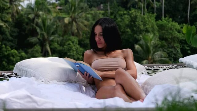 a girl on a hammock in an expensive villa, resting with a book under the sun during her honeymoon. Portrait with elegant, pretty, beautiful girl with perfect skin she read the book at morning
