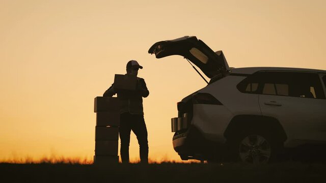 Parcel delivery concept. Male courier driver wearing protection mask and medical rubber gloves loads boxes during coronavirus. Man volunteer works stacks boxes in a opens delivery auto back door.