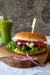 Freshly made burger on the cutting board on the wooden table and a green smoothie in the background, with copy space, vertical photo