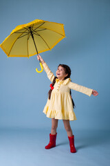 Happy cute little Girl Indian in red rubber boots, cotton dress holds in her hands yellow umbrella zones on a blue background in the studio. concept Spring, space for text - obrazy, fototapety, plakaty