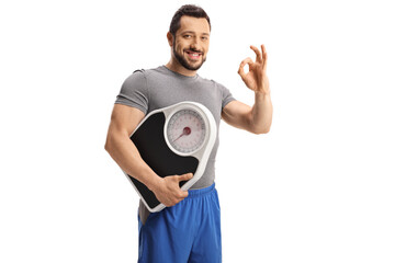 Smiling young man holding a weight scale and gesturing ok sign