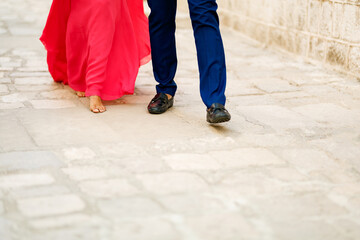 Barefoot bride in a long bright pink dress and the groom walking along a cobbled road, close-up