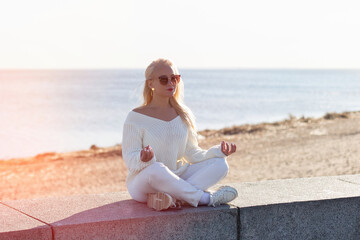 Fototapeta na wymiar Young beautiful blonde woman sitting near the sea and meditating at sunrise time