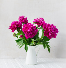 Purple garden peonies in a white enamel jug on a white wooden background, rustic style