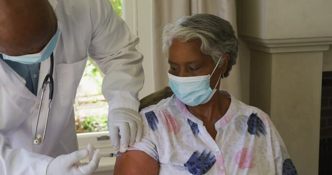 African american senior male doctor injecting covid-19 vaccine into african american woman at home