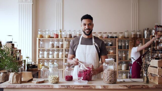Young man filling jar by dryed raspberries in zero waste shop, looking at camera.