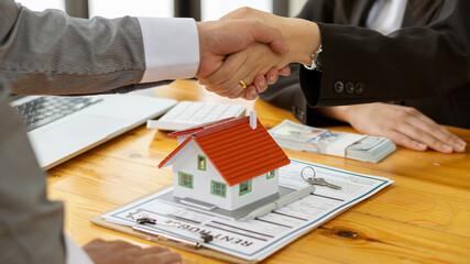 A young businessman shakes hands with a partner after Zen, a real estate investment contract document on a table with a house design and documents and money and a laptop.