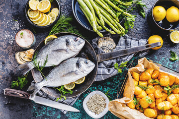 Fresh uncooked dorado or sea bream fish with lemon ,asparagus, herbs, olive oil and spices over black backdrop, top view
