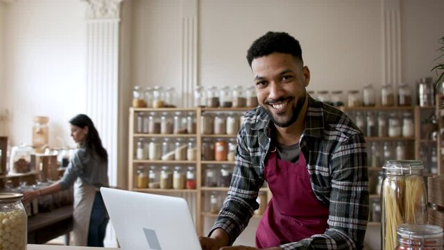 Owner sitting in front of laptop and looking at camera in zero waste shop.