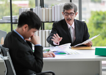 Elderly Asian businessman leader in a formal suit sitting at table and pointing the finger at the employee's face, and blaming him for the errors. Employee in foreground with bookshelf in background