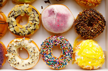 Top view flat lay of a colorful group of twelve delicious donuts in a white paper box on a wooden table. Collection of dozen donuts with glazes, sprinkles, almonds, peanuts, and chocolate chips