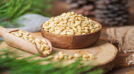 A plate and a scoop filled with pine nuts are on a wooden board on the table. Nearby are pine branches, gray fabric. 