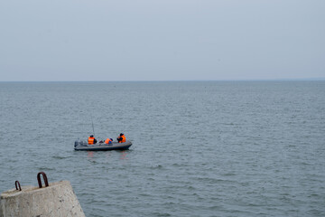 Three fishermen with rods dressed in protective orange vests sitting with backs to viewer in black rubber boat fishing near shore on background of sea horizon, on left can see top of breakwater.