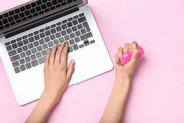 Woman squeezing stress ball while working with laptop on color background