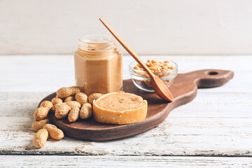 Jar with tasty peanut butter, bread and nuts on light wooden background