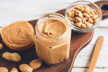 Jar with tasty peanut butter, bread and nuts on light wooden background