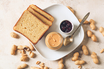 Jar with tasty peanut butter, toasts and jam on light background